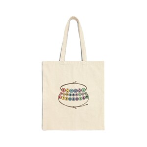 May include: A natural-coloured canvas tote bag featuring a graphic of two brown string bracelets with colourful beads. The top bracelet spells SCHOOL with small icons, and the bottom bracelet spells LIBRARIAN with additional small icons.