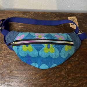 May include: A bum bag featuring a blue, green, and purple floral pattern on the front panel, accented by denim side panels and a blue adjustable strap. The bag includes a black zip with a multi-coloured iridescent metallic finish.