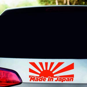 Puede incluir: Una pegatina roja y blanca en la parte trasera de un coche. La pegatina presenta un diseño de sol naciente con el texto "Made In Japan" en rojo.