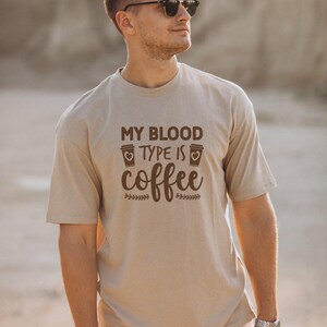 Puede incluir: Una camiseta de manga corta de color beige con el texto marrón "MY BLOOD TYPE IS coffee" centrado en el pecho. El diseño incluye dos pequeños iconos de tazas de café con corazones y detalles decorativos de hojas junto a la palabra coffee.