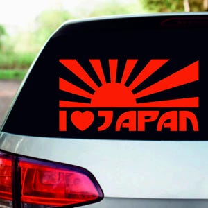 May include: Red "I Heart Japan" sticker on a car window. The design features a rising sun with rays and the text "I ♥ JAPAN" in red. The sticker is applied to the rear window of a silver car.