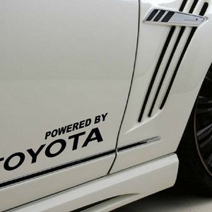 Może przedstawiać: Biały samochód z czarnymi listwami ozdobnymi i czarną naklejką z napisem "Powered by Toyota" na boku.