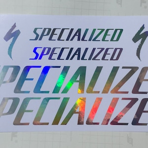May include: A sheet of iridescent vinyl stickers with the word "Specialized" printed four times in a bold, slanted font. Two of the stickers feature the Specialized logo, a stylized lightning bolt.