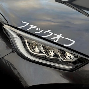 Könnte beinhalten: Nahaufnahme einer dunkelgrauen Motorhaube mit einem weißen Aufkleber. Der Aufkleber zeigt japanische Schriftzeichen. Der Autoscheinwerfer ist sichtbar, mit einem hellen, modernen Design. Der Lack des Autos reflektiert den Himmel.