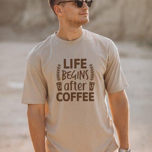 Puede incluir: Una camiseta de manga corta de color beige con el texto en marrón oscuro LIFE BEGINS after COFFEE, flanqueado por dos gráficos de ramas con hojas verticales y dos pequeños iconos de tazas de café con corazones en el frente.