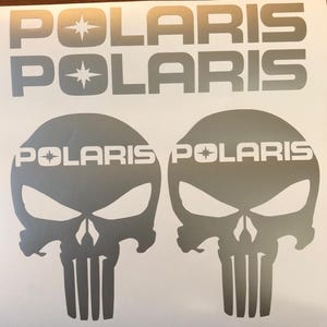 May include: Two silver skull decals with the word "POLARIS" printed in white on each skull. The skulls have a star symbol next to the word "POLARIS".