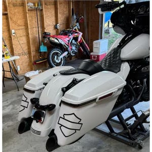 Könnte beinhalten: Ein weißes Motorrad mit schwarzen Akzenten und einem schwarzen Sitz steht in einer Garage. Das Motorrad hat ein Schildlogo an der Seite und zwei Auspuffrohre. Im Hintergrund ist ein rotes Dirtbike zu sehen.