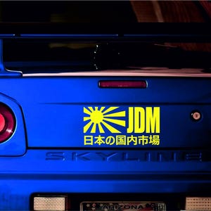Può includere: Primo piano della parte posteriore di un'auto blu con un adesivo JDM giallo e nero. L'adesivo presenta un design con sole nascente e testo giapponese. L'auto ha fanali posteriori rossi e uno spoiler.