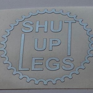 May include: A silver sticker with a white circle and the text "SHUT UP LEGS" in a decorative font.