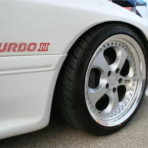 Puede incluir: Coche blanco con una llanta de aleación plateada y un neumático negro. El coche tiene el texto "TURBO II" en rojo en el lateral. El coche está estacionado sobre una superficie pavimentada.