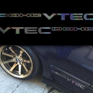 May include: Holographic "DOHC VTEC" car decals. The decals are in a multi-colored, iridescent finish. The image also shows a gold-colored car wheel and a section of a dark blue car with a matching decal.