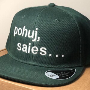 Può includere: Un cappello da baseball verde scuro con una frase ricamata in bianco "pohuj, saies..." sul davanti. Il cappello ha una visiera piatta e un'etichetta bianca "FLAT VISOR" sul lato inferiore della visiera.