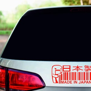 Pode incluir: Adesivo vermelho "Made in Japan" na janela de um carro. O adesivo apresenta um personagem de desenho animado, texto japonês e um design de código de barras. A luz traseira do carro é visível.