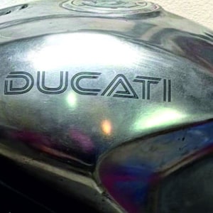 Puede incluir: Primer plano de un depósito de combustible de motocicleta con la palabra "DUCATI" en letras negras. El depósito tiene un acabado plateado metálico con algo de desgaste. La imagen está bien iluminada, destacando la forma del depósito y el logotipo de la marca.