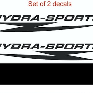 Può includere: Due decalcomanie nere con la scritta "HYDRA-SPORTS" in un carattere audace e stilizzato. Ogni decalcomania ha un design a freccia nera sotto il testo. Le parole "Set of 2 decals" sono in rosso in alto.