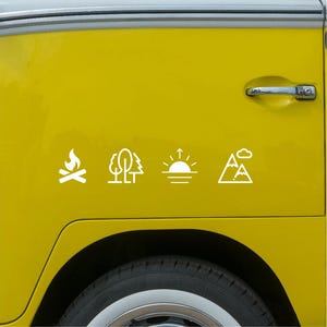 Könnte beinhalten: Weiße Camping-Themenaufkleber auf einem gelben Fahrzeug. Die Aufkleber zeigen ein Lagerfeuer, Bäume, einen Sonnenuntergang und einen Berg mit einer Wolke. Die Aufkleber sind horizontal an der Seite des Fahrzeugs angeordnet.
