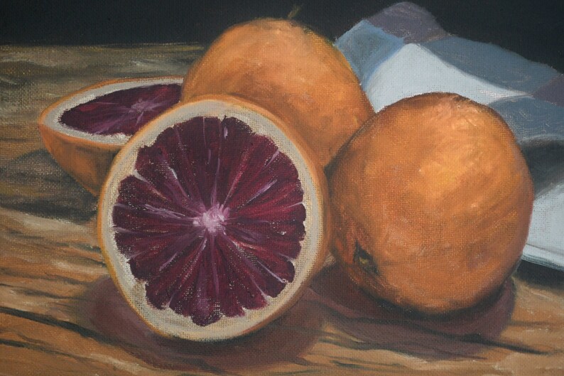 K&ouml;nnte beinhalten: Ein Stillleben mit drei Orangen, von denen zwei halbiert sind und ihr tiefrotes Fruchtfleisch zeigen. Die Orangen sind auf einem Holztisch mit einem blau-wei&szlig;en Tuch im Hintergrund angeordnet.