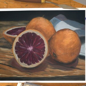 K&ouml;nnte beinhalten: Ein Stillleben mit drei Orangen, von denen zwei ganz und eine halbiert ist, wodurch das rote Fruchtfleisch sichtbar wird. Die Orangen liegen auf einem Holztisch mit einem blau-wei&szlig;en Tuch im Hintergrund.