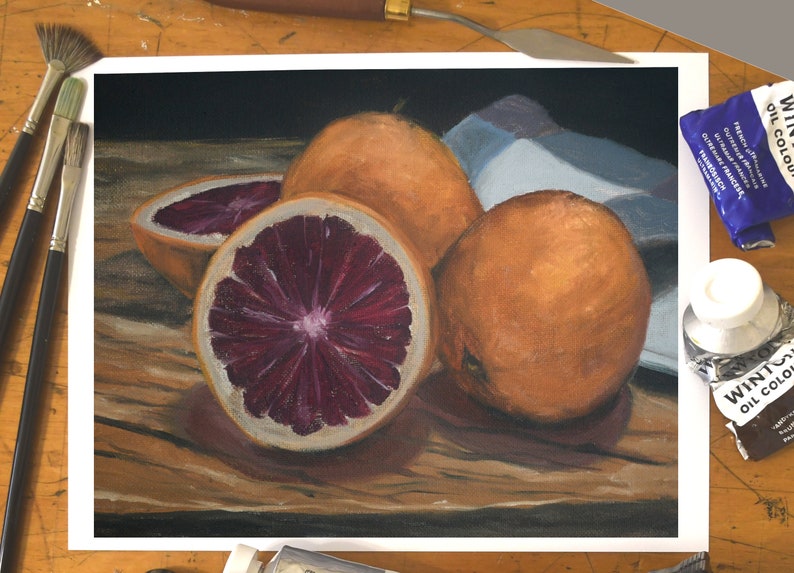 K&ouml;nnte beinhalten: Ein Stillleben mit drei Orangen, von denen zwei ganz und eine halbiert ist, wodurch das rote Fruchtfleisch sichtbar wird. Die Orangen liegen auf einem Holztisch mit einem blau-wei&szlig;en Tuch im Hintergrund.