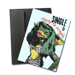 May include: A greeting card with a green creature illustration wearing a leopard print bikini and long green hair. The card reads "SINGLE & ready to mingle" in black and red text. The card is next to a black envelope.