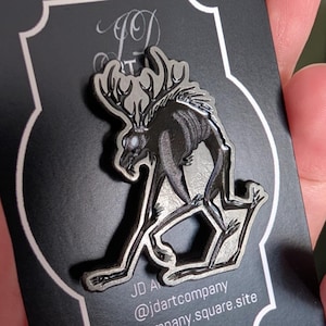 Peut inclure: Épingle en métal argenté représentant une créature squelettique avec des bois, en position accroupie. L'épingle est présentée sur une carte noire avec le texte "JD Art Company" et une adresse de site web.