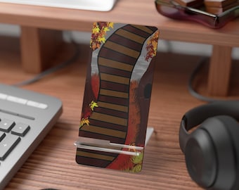 May include: A phone stand with a fall-themed design. The stand features a brown wooden bridge with autumn leaves and foliage. The stand is made of clear acrylic and is designed to hold a phone or tablet.