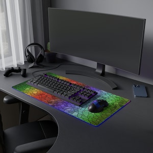 May include: A gaming setup featuring a black keyboard, mouse, and a colorful, mosaic-patterned mousepad. A large monitor, headphones, and a game controller are also visible. The desk is dark, and the overall aesthetic is modern and tech-focused.