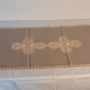 Könnte beinhalten: Ein beigefarbener gehäkelter Tischläufer mit einem zarten Blumenmuster. Der Läufer ist aus einem weichen, leichten Material gefertigt und hat einen schlichten, eleganten Stil.