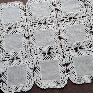 May include: White crocheted tablecloth with a square pattern. The tablecloth has a scalloped edge and is made of delicate lace.