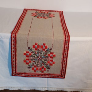 Könnte beinhalten: Ein beigefarbener Tischläufer aus Leinen mit roten, schwarzen und weißen gestickten Blumendesigns. Der Läufer hat einen geometrischen Musterbord.