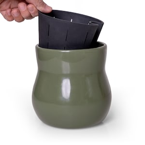 May include: A glossy olive green ceramic planter with a rounded base and a wide opening. A removable black plastic inner liner with vertical drainage slits is partially lifted out of the top of the ceramic pot, revealing its functional design.