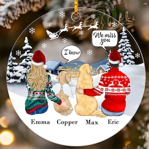 May include: A clear, round Christmas ornament with a winter scene. It features two people and two dogs, all wearing Santa hats. Text bubbles say "I know" and "We miss you." The names Emma, Copper, Max, and Eric are printed below.