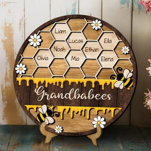 May include: A wooden honey comb shaped sign with the words "Grandbabees" and the names Liam, Lucas, Noah, Olivia, Mia, Ethan, Elena and Ella. The sign is decorated with yellow honey dripping down the sides and bees.