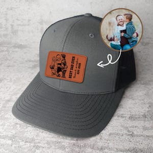 May include: A gray trucker hat with a black mesh back and a curved brim. The hat features a rectangular brown leather patch with a family photo and the text "BEST DAD EVER". A circular photo of a family is also included.