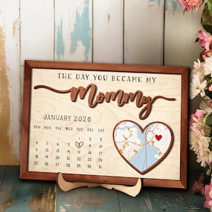 May include: Wooden framed sign with the text "THE DAY YOU BECAME MY Mommy" and a calendar for January 2026. A heart-shaped map with a red heart is on the right. The sign is supported by a wooden stand.