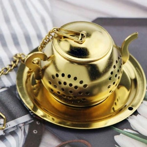 May include: Gold-toned metal tea infuser shaped like a teapot with a lid and a chain. The infuser sits on a matching gold-toned metal saucer.