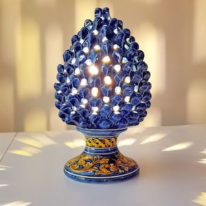 Peut inclure: Une lampe en céramique bleue et jaune en forme de pomme de pin avec des détails complexes. La lampe a une base avec un motif floral.
