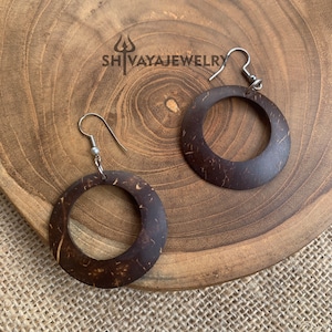 Puede incluir: Un par de pendientes de aro de cáscara de coco marrón con ganchos plateados. Los pendientes están sobre una superficie de madera.