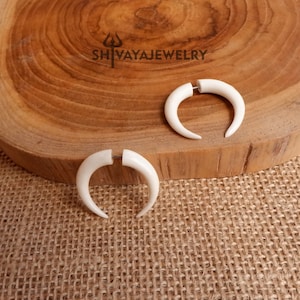 May include: A pair of white horn-shaped earrings. The earrings are made of bone and have a silver-coloured post. The earrings are on a wooden surface.
