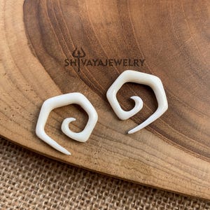 Puede incluir: Dos expansores de oreja en espiral blancos, hechos de hueso, sobre una superficie de madera.