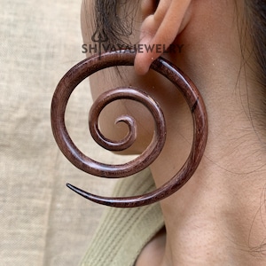 May include: A dark brown wooden spiral ear gauge. The gauge is a single piece of wood that is carved into a spiral shape. The gauge is worn in the ear of a person.