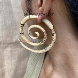 May include: A large, spiral-shaped wooden earring with a natural wood grain pattern and dark brown streaks. The earring is worn in the upper lobe of a person's ear.