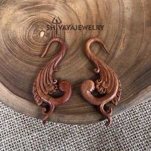 May include: A pair of wooden earrings with a detailed, intricate design. The earrings are shaped like a bird with a curved beak and a long, flowing tail. The earrings are a rich brown color and have a natural, rustic finish.
