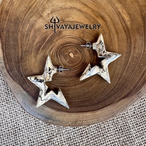 May include: A pair of star-shaped earrings made of light-colored wood with dark gray and black veining. The earrings are on a wooden surface with the text "SHIVAYAJEWELRY" in the upper center.