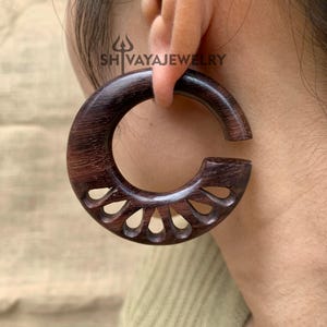 May include: Dark brown wooden hoop earring with a carved floral design. The earring is a large, circular shape with a smooth, polished finish. The text "SH VAYAJEWELRY" is visible on the earring.