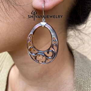 May include: Oval earrings with a silver hook and an openwork design. The earrings have a pearlescent, iridescent material with a silver border and swirling patterns. The earrings are displayed on a person's ear.