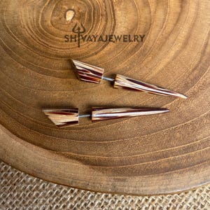May include: A pair of wooden earrings with a geometric design. The earrings have a light brown and dark brown striped pattern and a silver post. The earrings are on a wooden surface with the text "SHIVAYAJEWELRY".