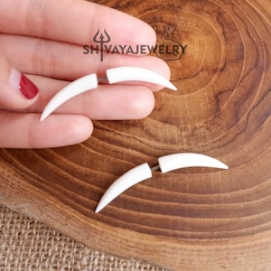 Può includere: Un paio di orecchini a forma di zanna bianca finta. Gli orecchini sono realizzati in un materiale liscio e bianco e hanno una forma curva. Gli orecchini sono mostrati su una superficie di legno.