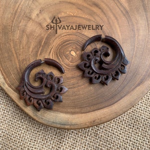 May include: A pair of dark brown wooden earrings featuring a carved floral design and a spiral shape. The earrings are presented on a wooden surface, with the text "SHIVAYAJWELRY". These earrings have a distinctive, bohemian style.