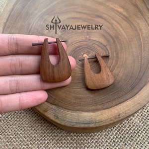 Könnte beinhalten: Ein Paar Holzohrringe mit einer einzigartigen, asymmetrischen Form. Die Ohrringe sind aus dunkelbraunem Holz gefertigt und haben ein schlichtes, minimalistisches Design. Die Ohrringe werden auf einer Holzoberfläche gezeigt.